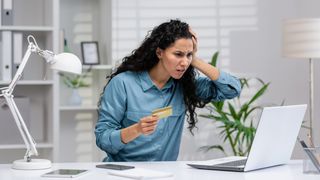 Woman sat at an office desk in front of a laptop holding a credit card looking stressed
