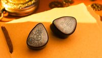 A pair of Dunu DaVinci Vitruvian earbuds, with sepia faceplates etched with clockwork gear drawings, sits on a dark wooden desk