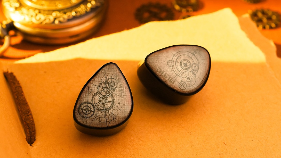 A pair of Dunu DaVinci Vitruvian earbuds, with sepia faceplates etched with clockwork gear drawings, sits on a dark wooden desk