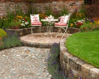 Small circle circular stone paved patio and lawn with a table and chairs seating with cobble cobbled pebble path and reclaimed brick raised bed