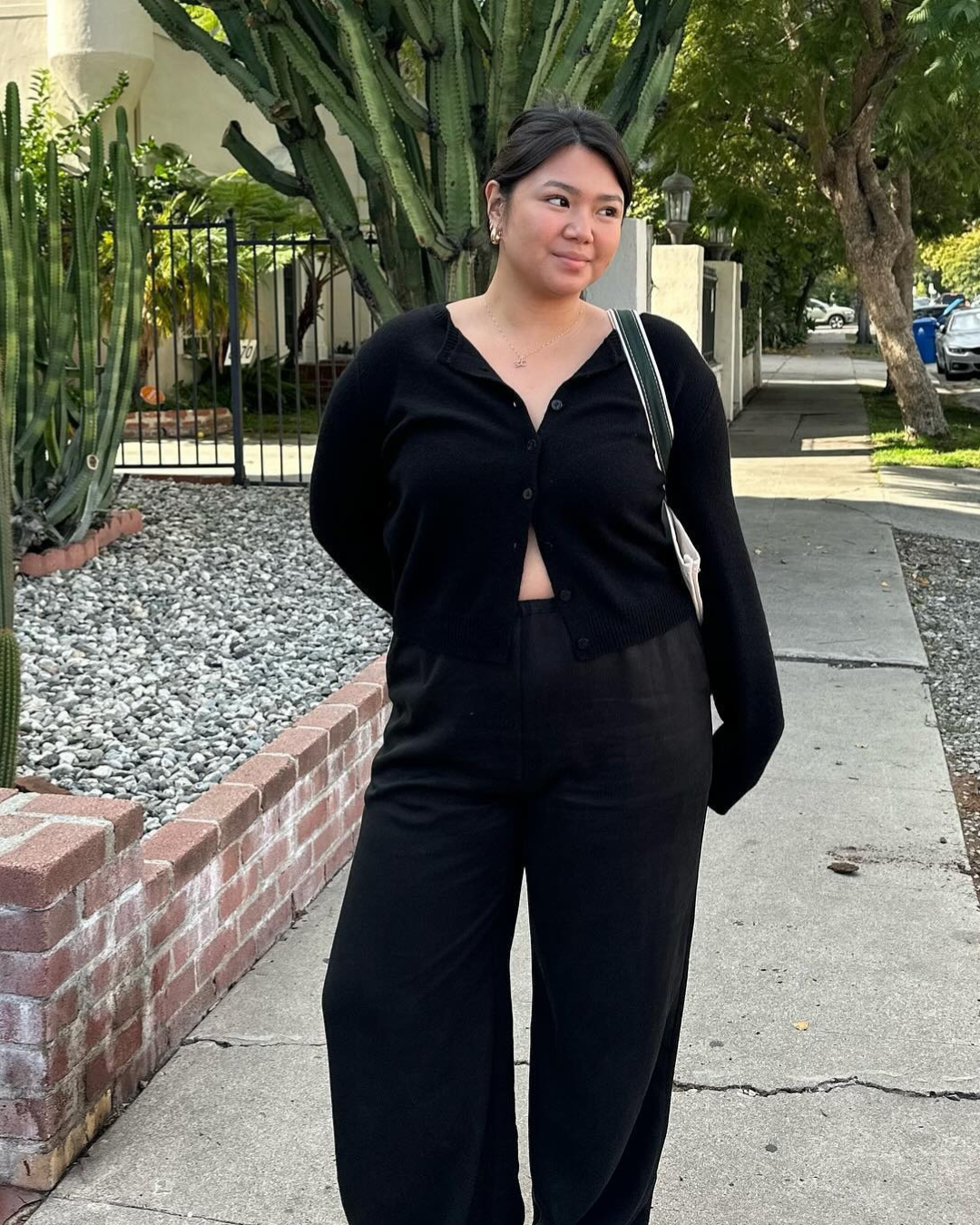 Influencer @marina_torres wears black satin trousers with black trainers and a partially unbuttoned black cardigan.