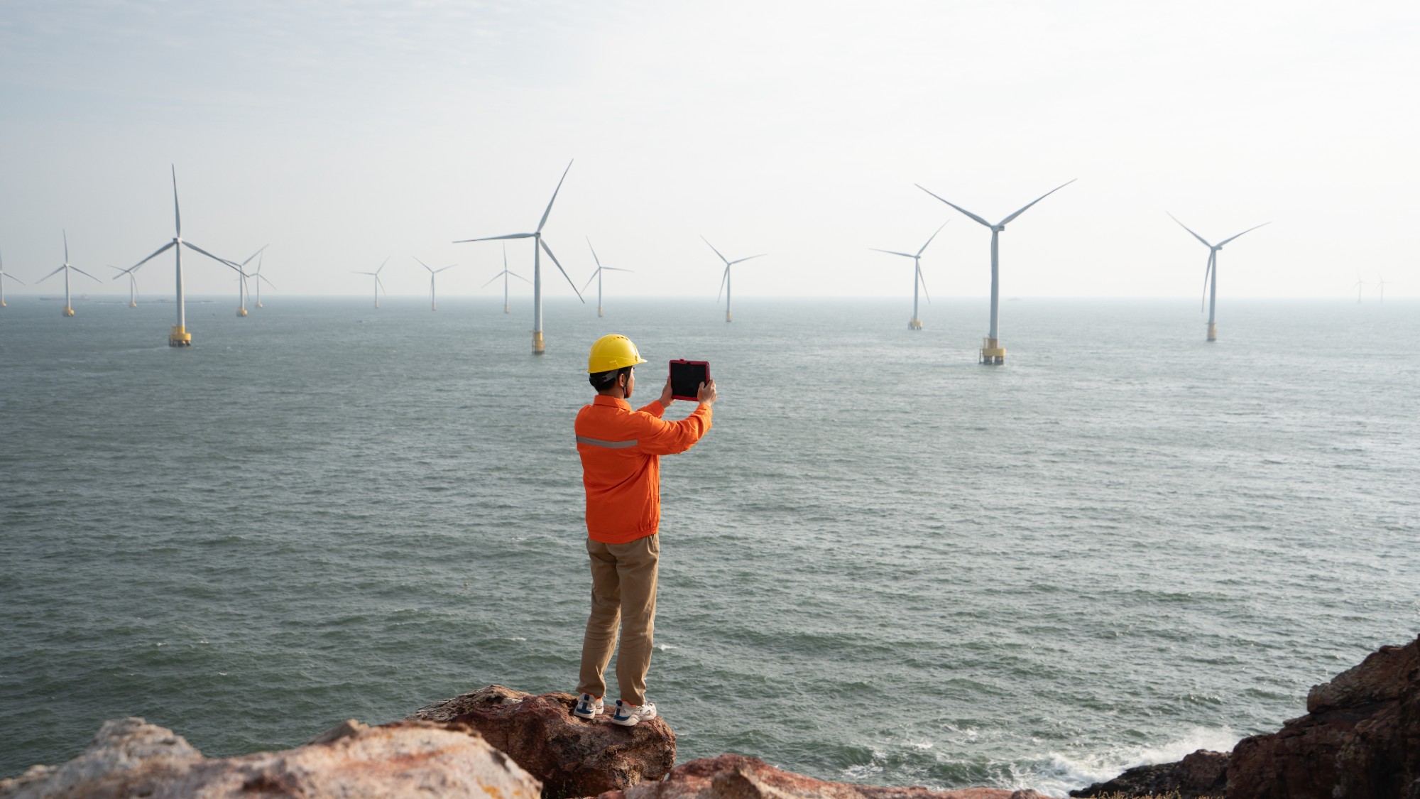 a man in orange construction gear takes a photo on a coastline. windmills are in the water offshore