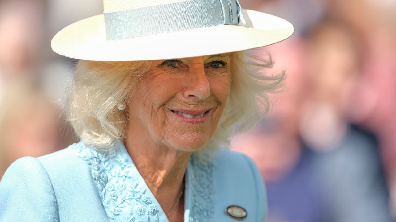 A shoulders-up photo of Queen Camilla wearing a blue coat and white hat with a blue ribbon and smiling