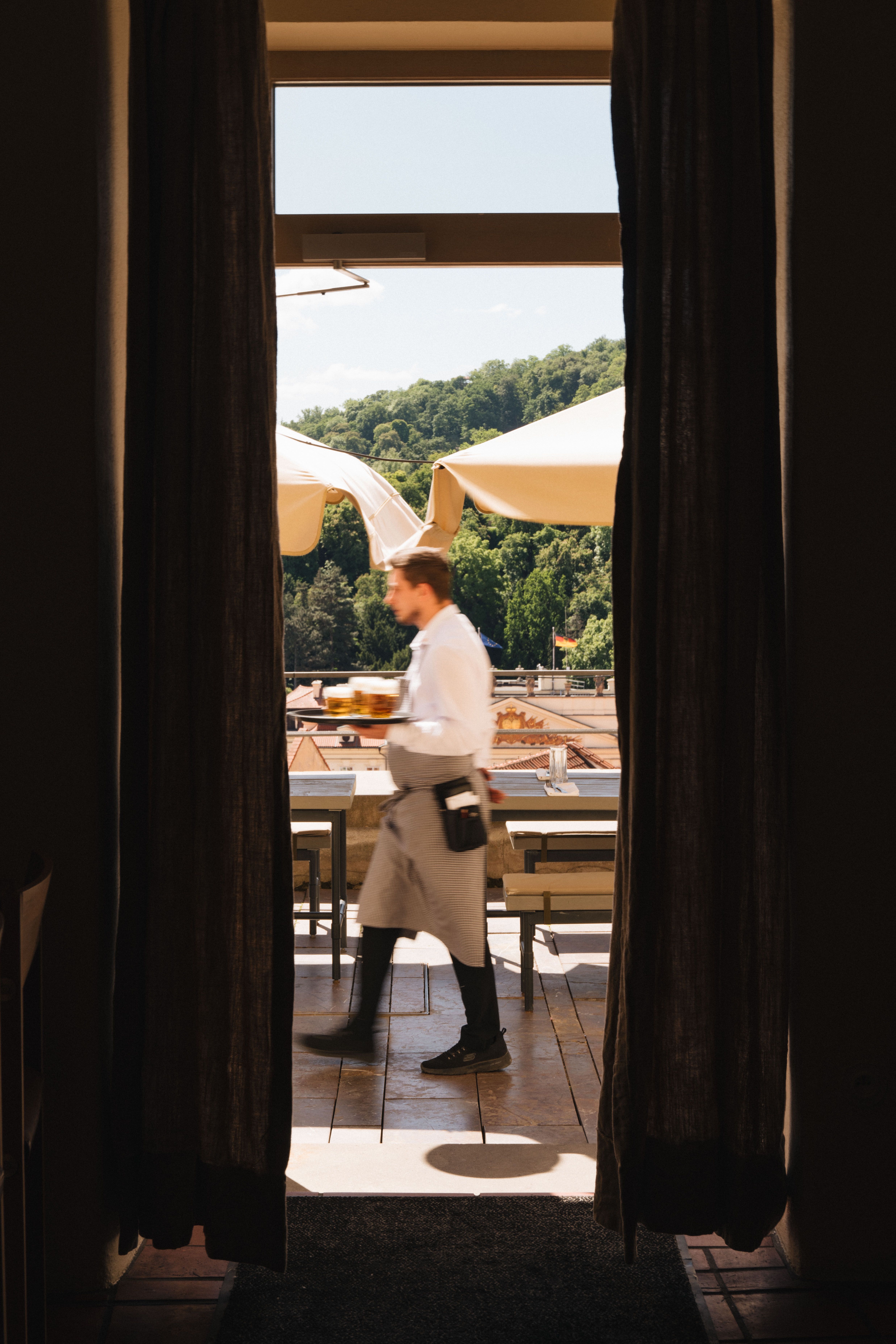 Two pictures of a restaurant, one caught from the inside of a sun-lit terrace, a waiter rushing by, and one of a plate of open sandwiches with fish, herbs, and olives, held by someone whose hand we see in front of a tiled water green wall.