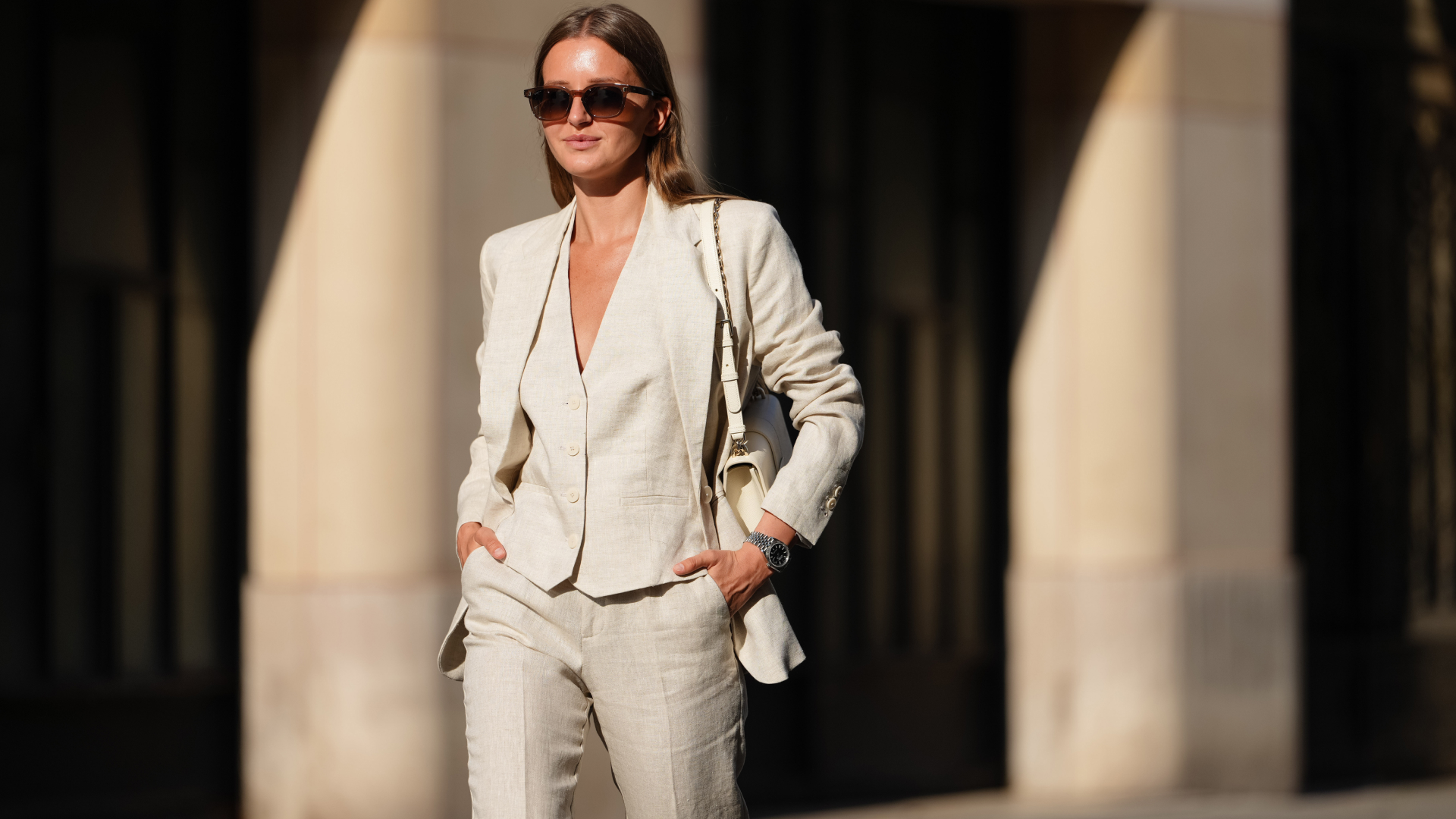 Diane Batoukina wears a light beige linen suit consisting of a single-breasted blazer with notched lapels and matching trousers. The blazer features a tailored fit with buttoned cuffs. Underneath, a matching waistcoat with a V-neckline and button fastening is visible. Sunglasses with dark lenses and a tortoiseshell frame are worn. A white leather shoulder bag with a gold chain strap is carried. A metallic watch adorns the wrist. Hair is styled straight and parted in the middle, during a street style fashion photo session, on May 14, 2025 in Paris, France
