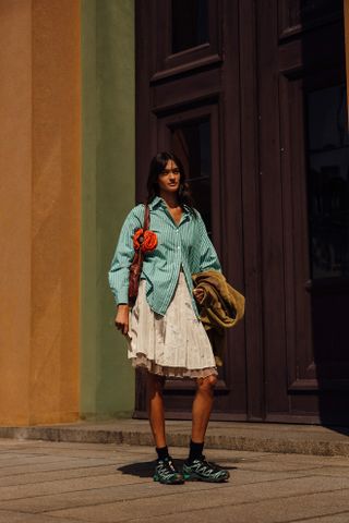 Woman at fashion week wearing preppy outfit featuring a green striped button down shirt, tan skirt, and black and green sneakers.