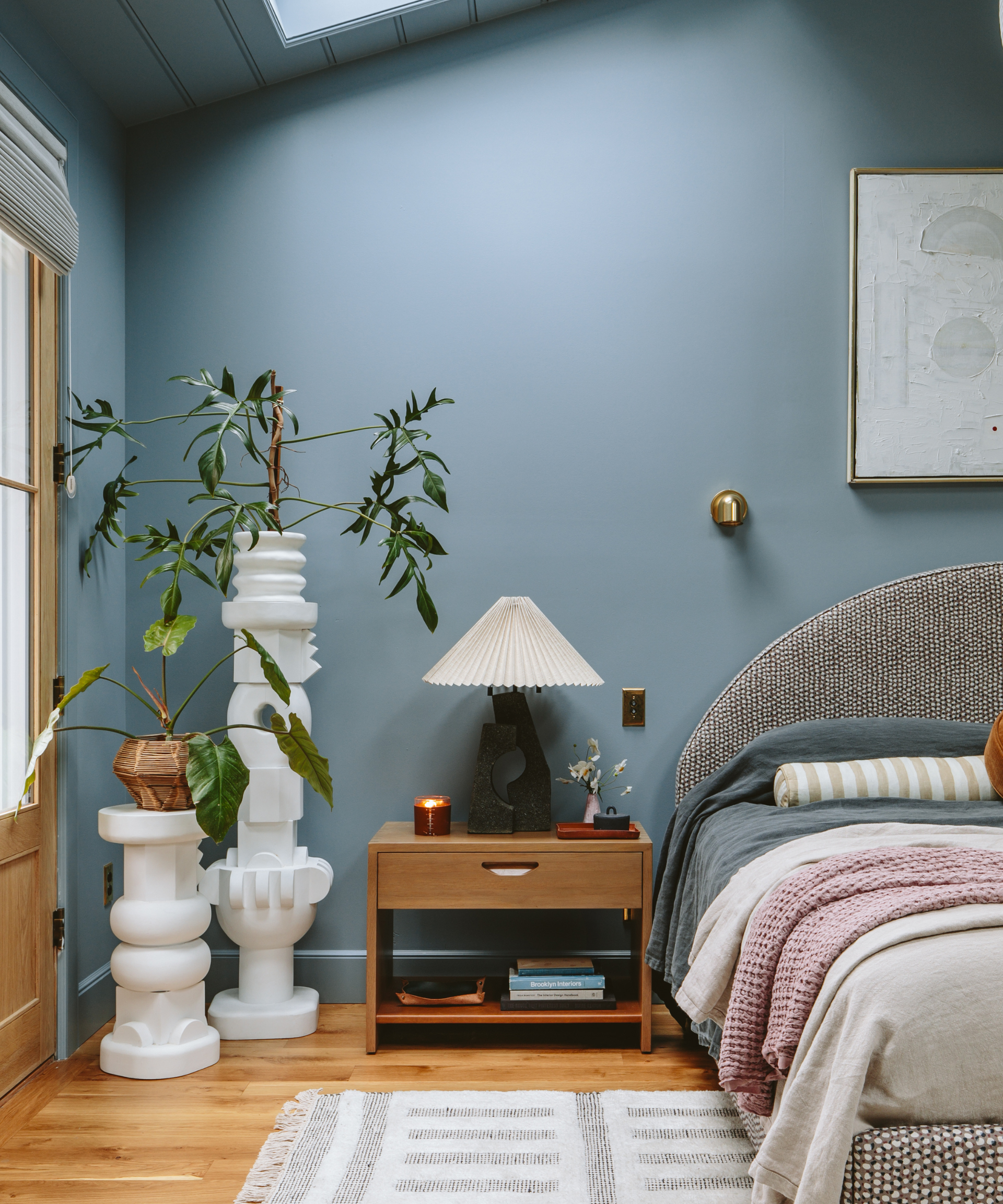A blue bedroom with a bed, side table and tall, structural, white plant holders.