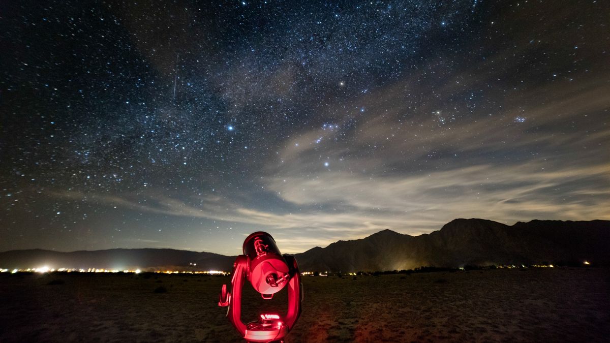 a telescope points upward to a starry sky in front of a mountain range