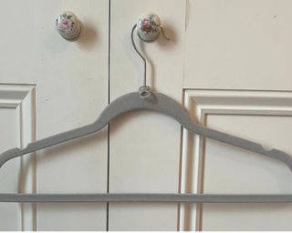 A gray velvet hanger with a pull tab on the neck hanging on a floral door knob on a white paneled cupboard door