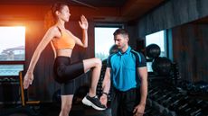 A woman performs step-ups in a gym, swinging her arms and raising her knee. Next to her a trainer is watching her form.