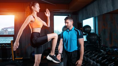 A woman performs step-ups in a gym, swinging her arms and raising her knee. Next to her a trainer is watching her form.