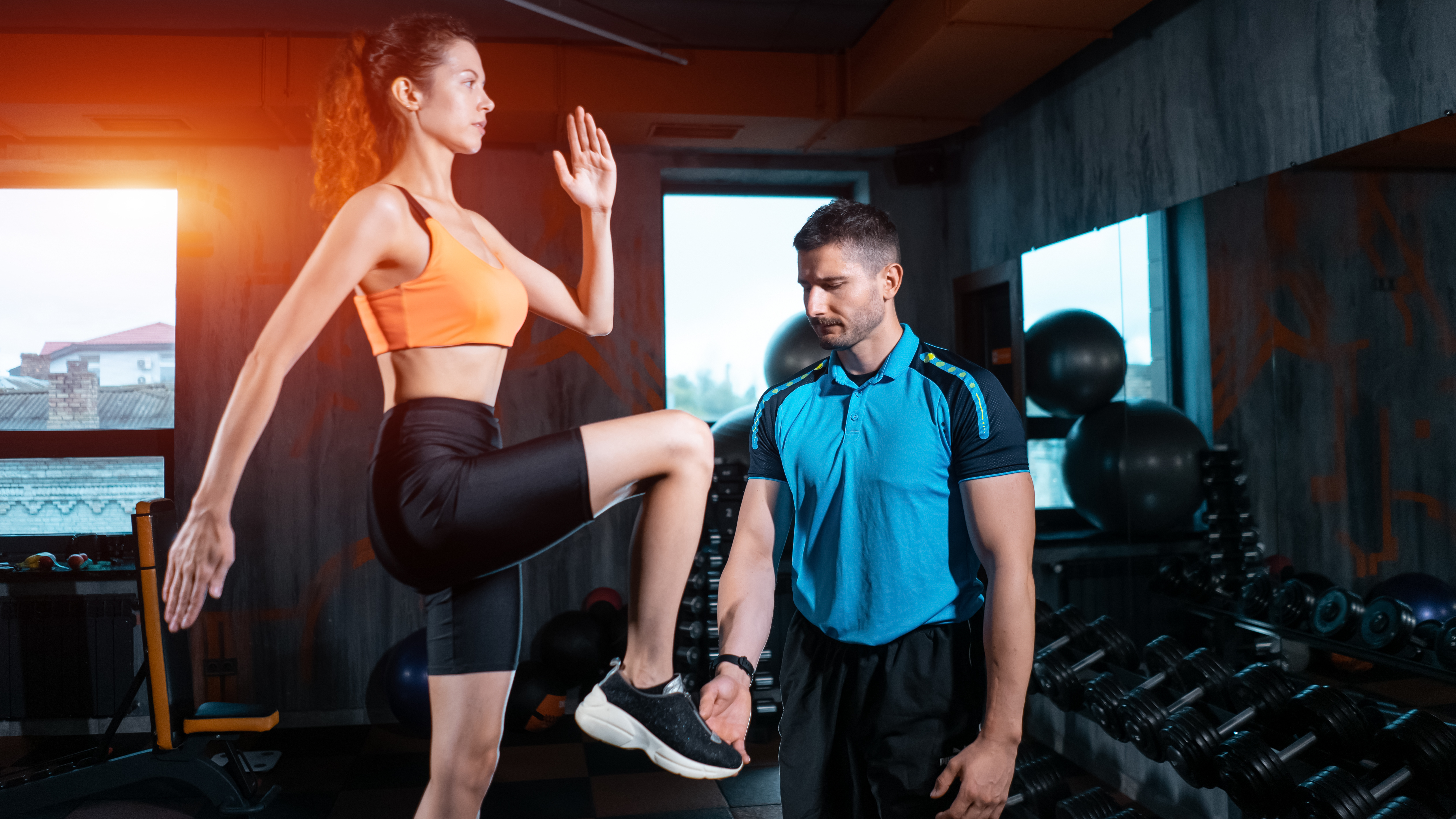 A woman performs step-ups in a gym, swinging her arms and raising her knee. Next to her a trainer is watching her form.