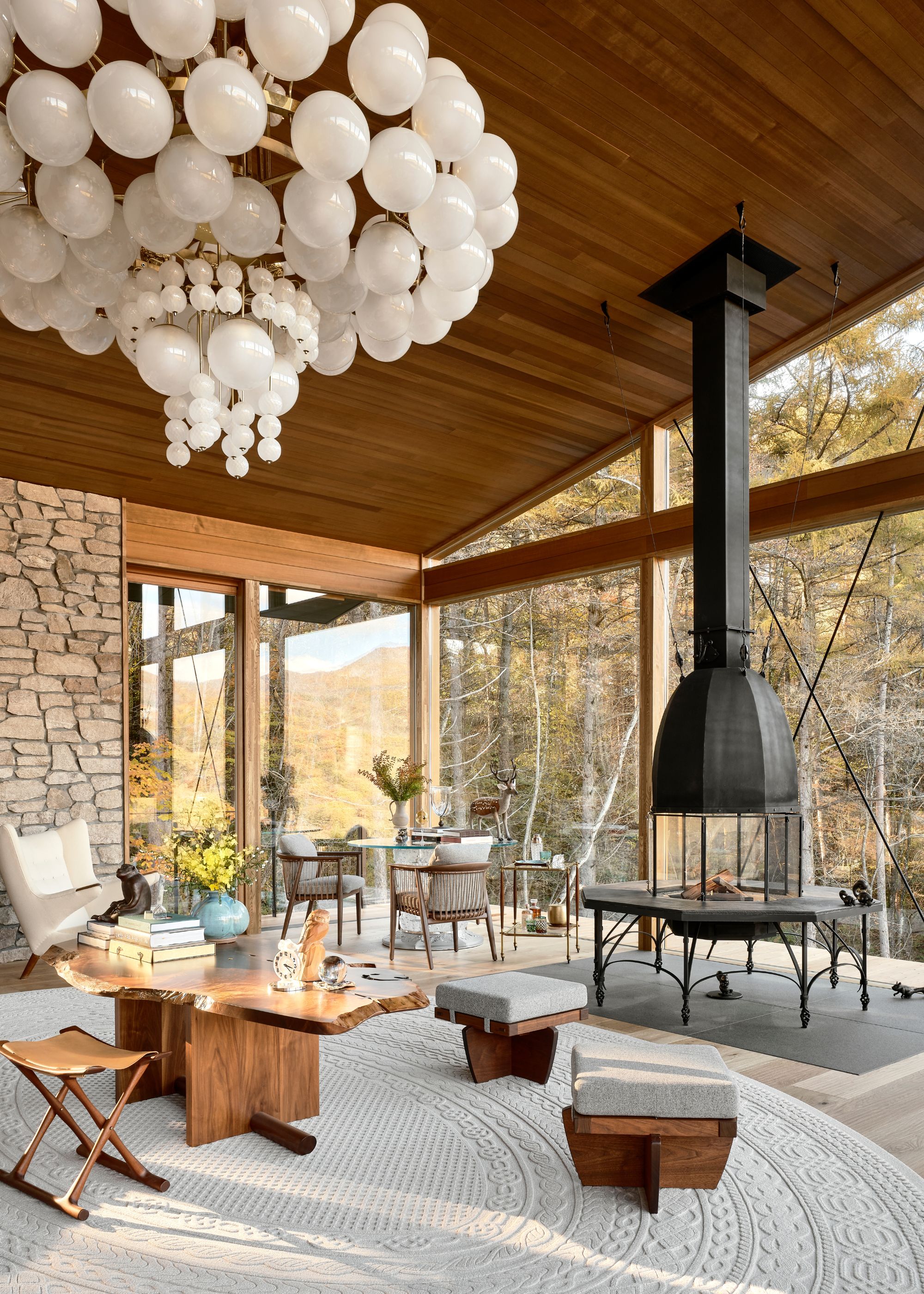 A Japanese living room with a wood paneled sloped ceiling, a large wood burner with chimney up to the ceiling buy the windows, and a large oval cream rug with a table and surrounding seating