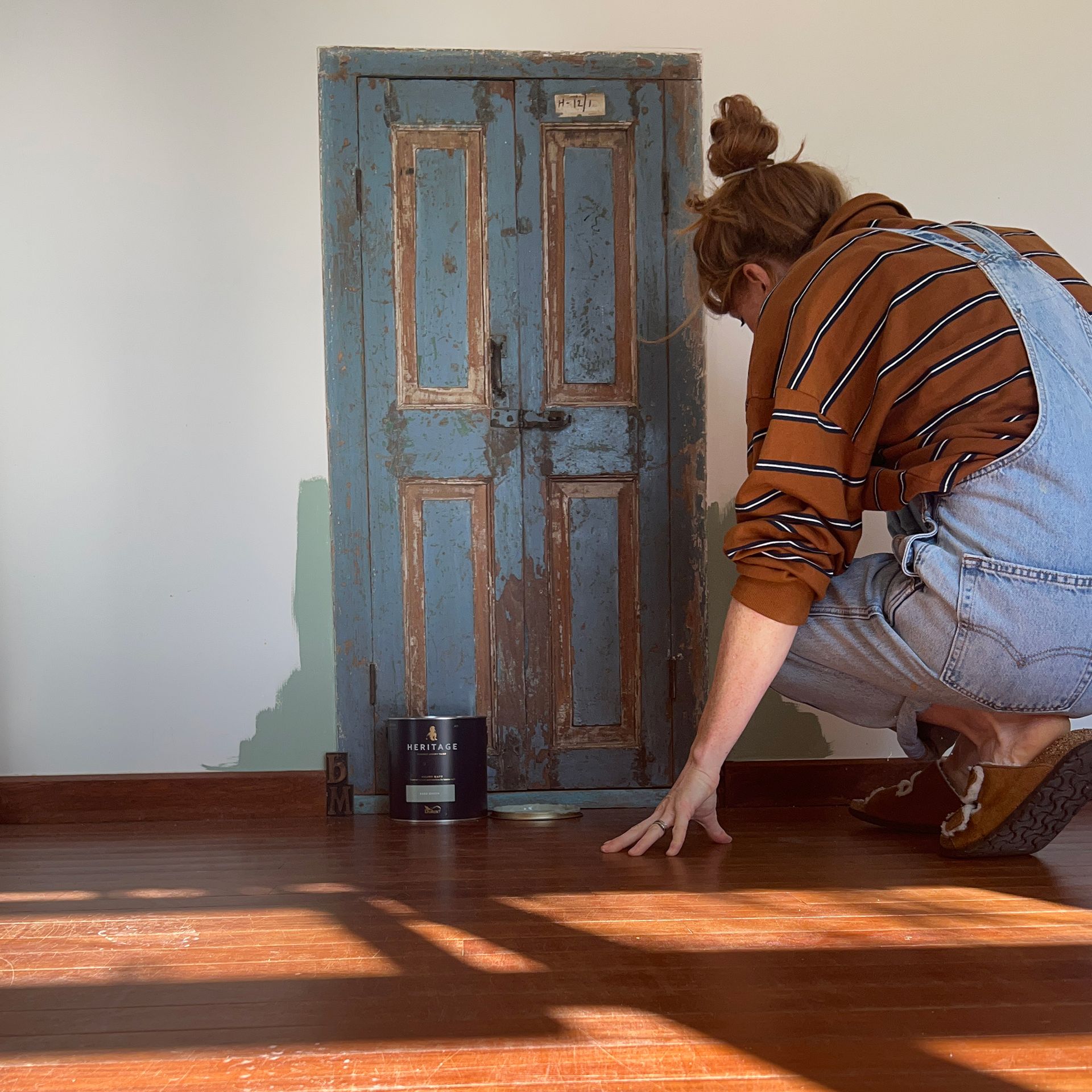 Woman crouching down painting neutral wall green around small blue door