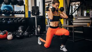 Woman performs lunge in gym holding dumbbells by her shoulders