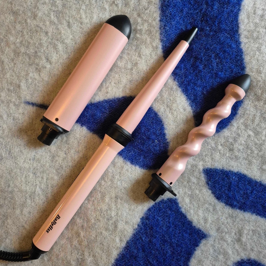 A picture of the BaByliss Curl and Wave Trio on a blue and cream blanket background. The curling tool comes with three interchangeable barrels: a conical wand, which creates defined curls, an oval wand, for loose mermaid-style waves and a twisted wand, for effortless, beach texture - all three are pictured here.