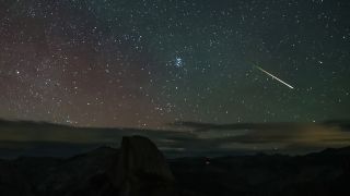 A bright meteor is pictured streaking across a starry night sky over a rocky landscape, as clouds lie low on the horizon.