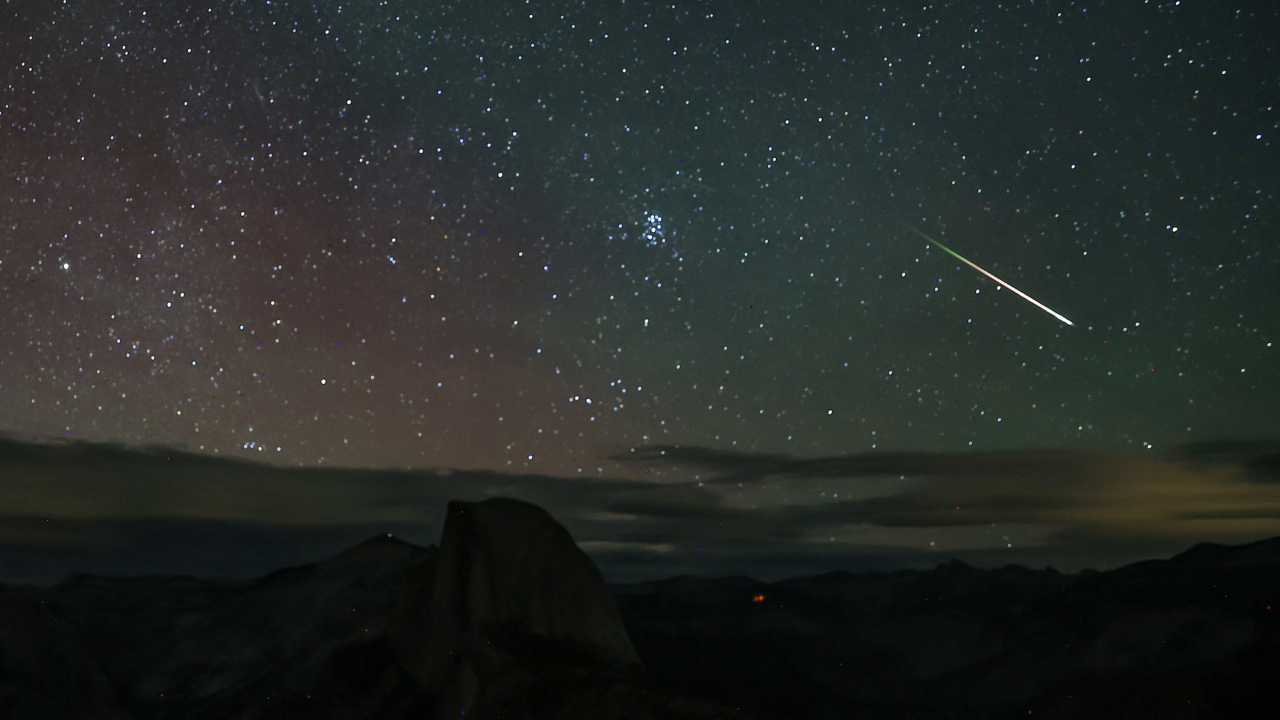 A bright meteor is pictured streaking across a starry night sky over a rocky landscape, as clouds lie low on the horizon.