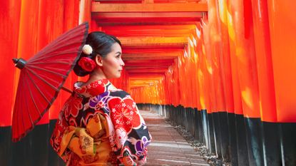 Woman wearing a patterned kimono and carrying a parasol. In the background are the red gates at Kyoto&amp;#039;s Fushimi Inari-taisha shrine.