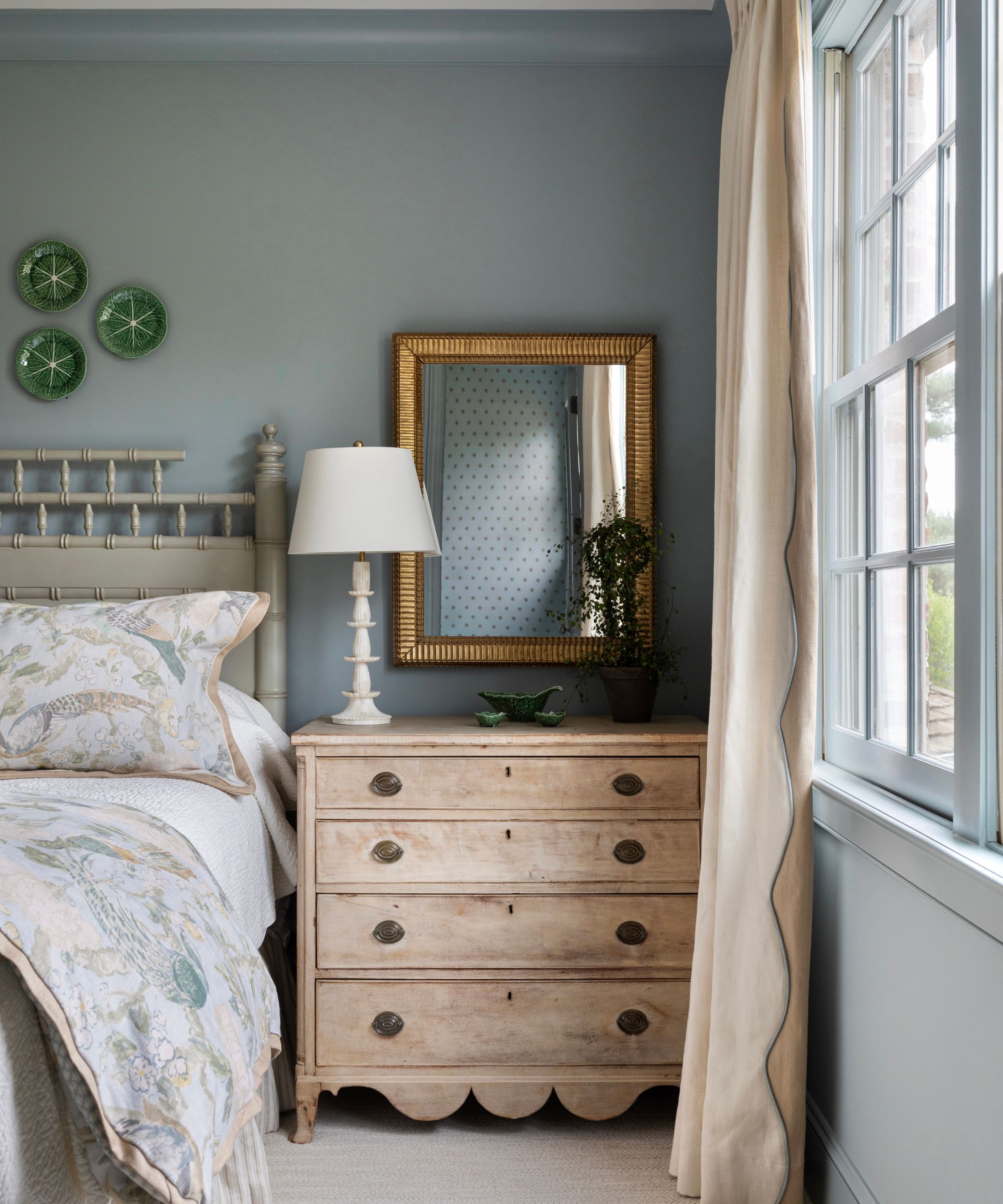 A transitional bedroom with blue-gray walls, a vintage wood dresser with a white lamp and gold mirror above it, soft blue patterned bedding, and light beige drapes.