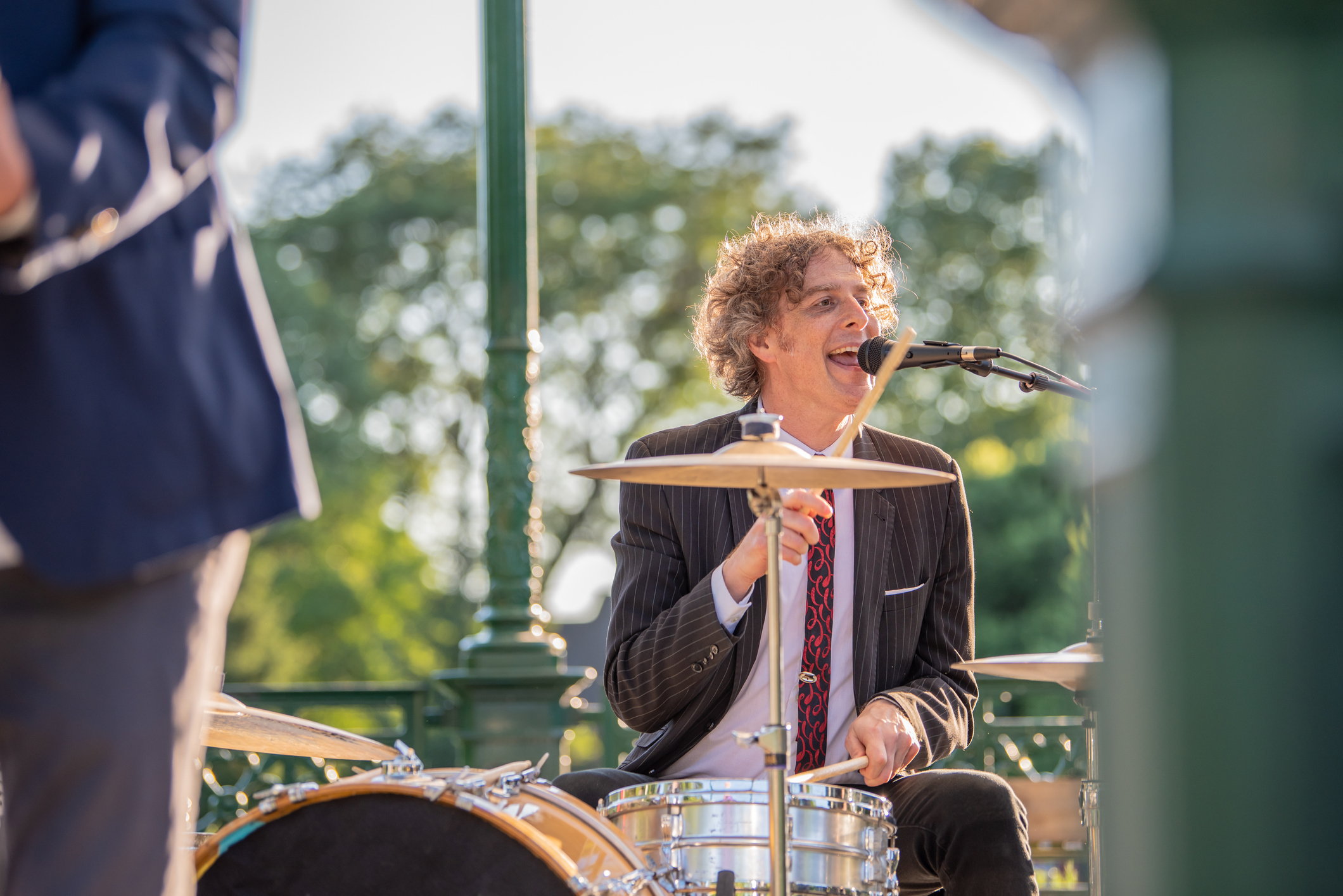 View just beyond the columns of a gazebo stage of an attractive forty something man playing drums in a band, outside in a park.