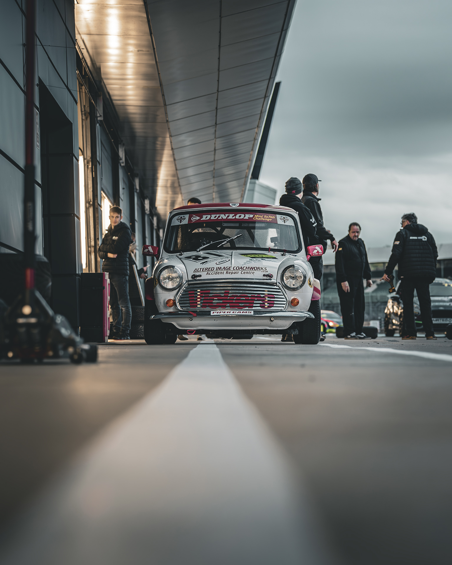 A classic Mini Cooper parked in a pit lane, surrounded by people preparing for a race under overcast skies