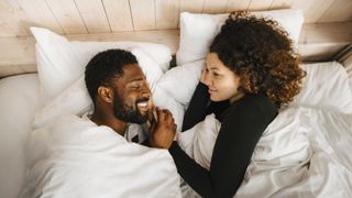 Man and woman lying in bed together under white sheets laughing
