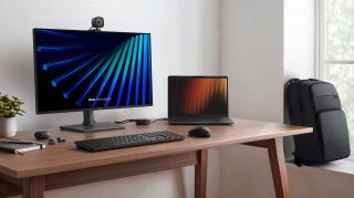 A modern workspace with a large monitor, Dell Pro laptop, keyboard, and mouse on a wooden desk, featuring a potted plant and a black backpack in the background.