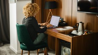 A woman with curly brown hair works at a desk in her hotel room