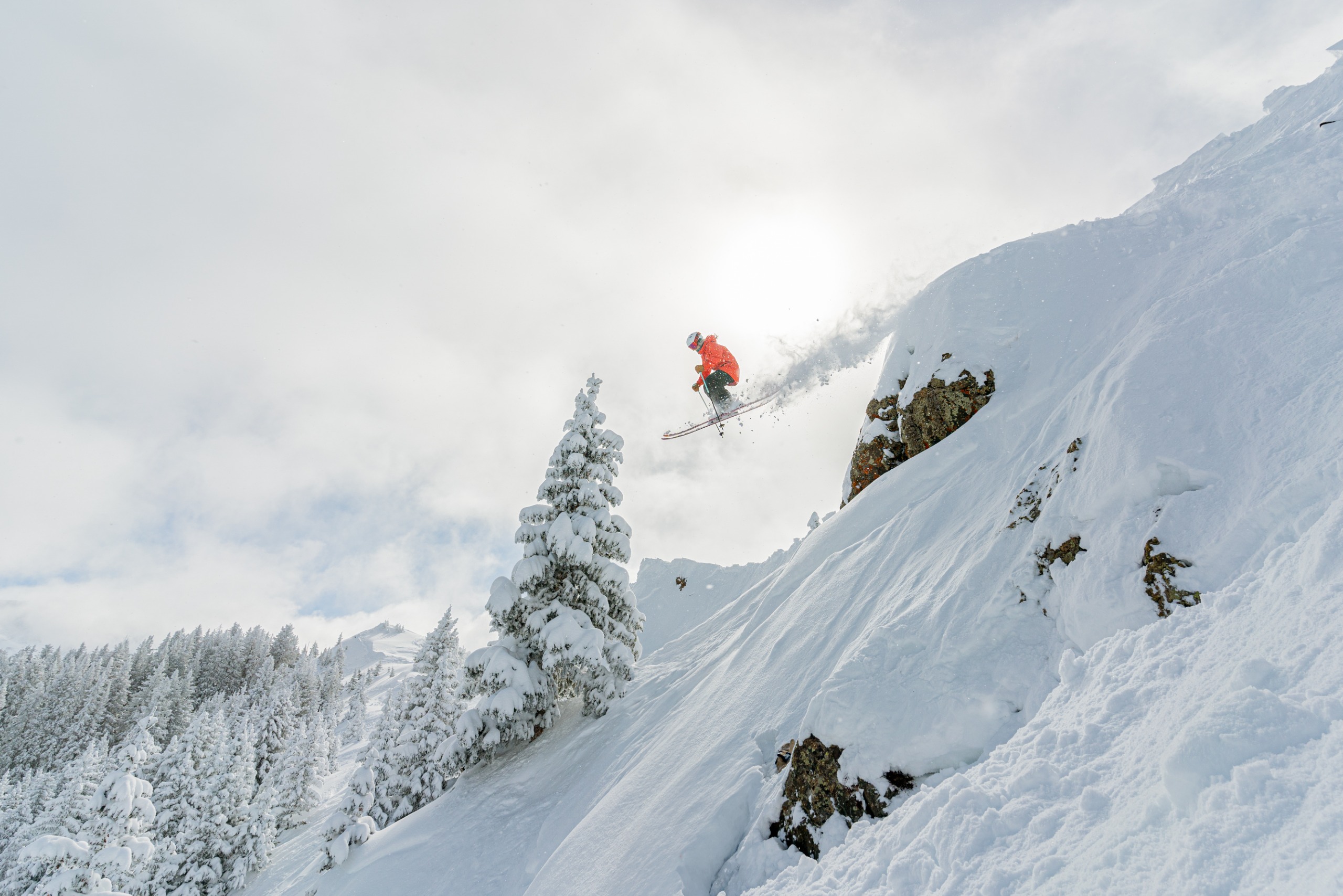 A skier in a red jacket and black trousers jumping off a snow-covered rock