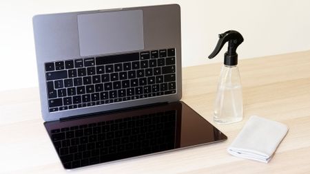 A photo of a MacBook flipped on its side, with a spray bottle and screen cleaning wipe next to it.