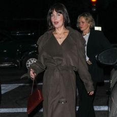 Dakota Johnson walks down the street wearing a dark brown belted coat with black knee-high boots.