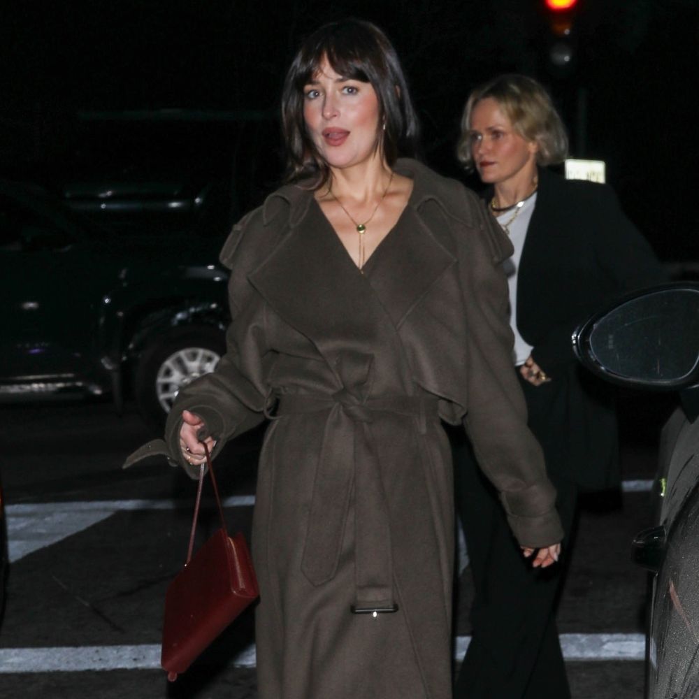 Dakota Johnson walks down the street wearing a dark brown belted coat with black knee-high boots.