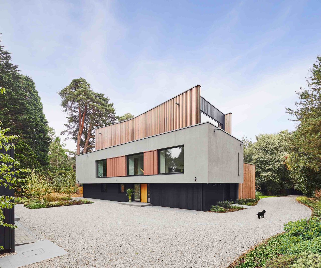 The front elevation of a modern self build, built by Baufritz, set in a woodland plot with an extensive gravel drive in front. The property features grey cladding and a timber-clad butterfly-style roof.
