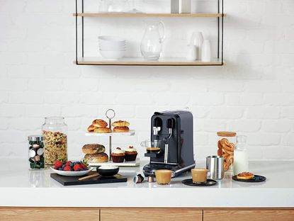 one of the best pod coffee machine options on a marble kitchen countertop, with cakes and baked good around it on a tiered trap, cups of coffee, a cannister of coffee pods, and a jar of granola