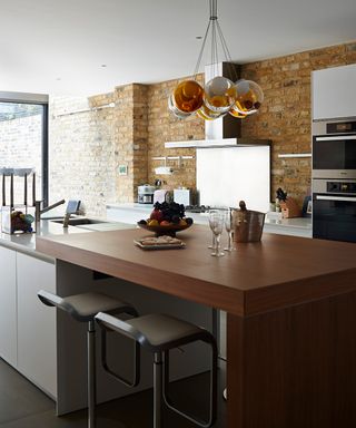 A close up shot of a wooden island with two bar stools in front of a metal splashback on an exposed brick wall