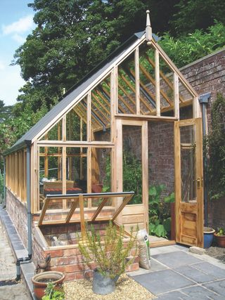 Wood-framed, Victorian-style greenhouse ideas shown leaning against a brick wall with slate paving leading in to the open door.