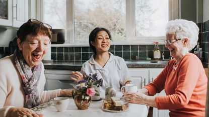 Three older women friends laugh together at a kitchen table.