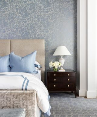 bedroom with blue patterned wallpaper, neutral bed and dark wood beside cabinet