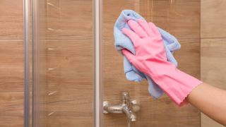 A gloved hand cleaning a glass shower wall with a microfiber cloth