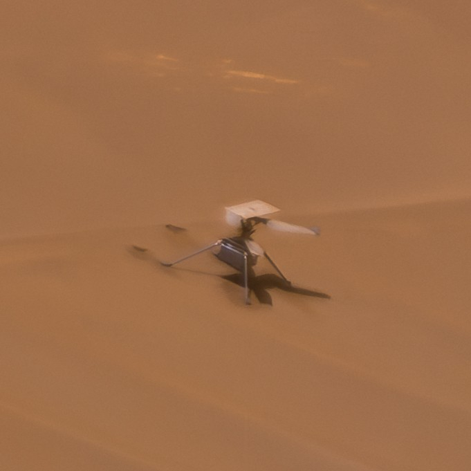 a four legged, double propeller helicopter drone sits on a sandy sloped hill, all rusted red in hue. the drone is missing part of a propeller.