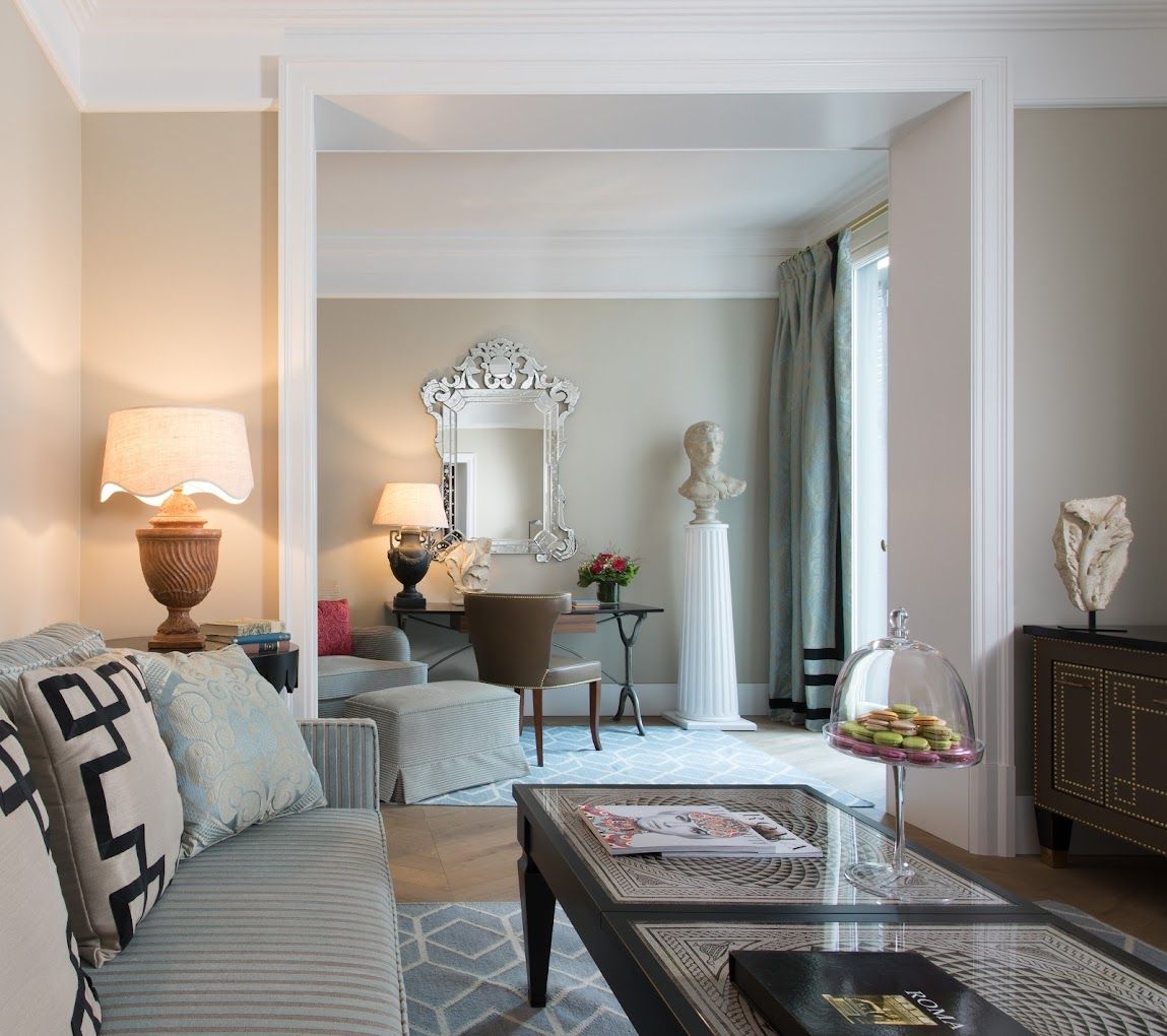 A neoclassic hotel room features print-filled upholstered furniture, marble sculptures inspired by Roman art, amphora-shaped glowing table lamps, and a neutral color scheme.