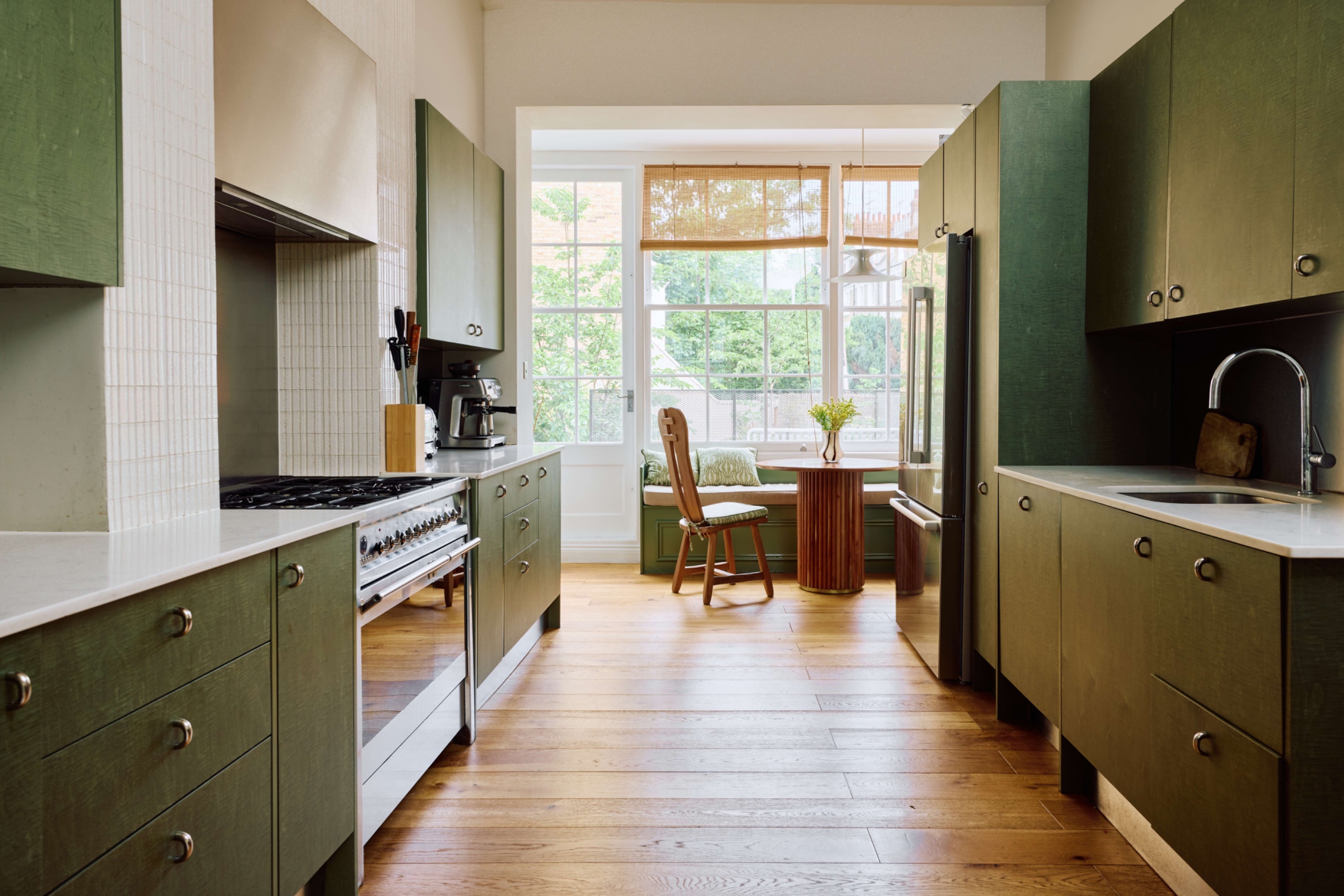 Green galley kitchen with pale countertops and wood flooring leading to a bay-window breakfast nook with built-in seating, a round wooden table and a wooden chair.
