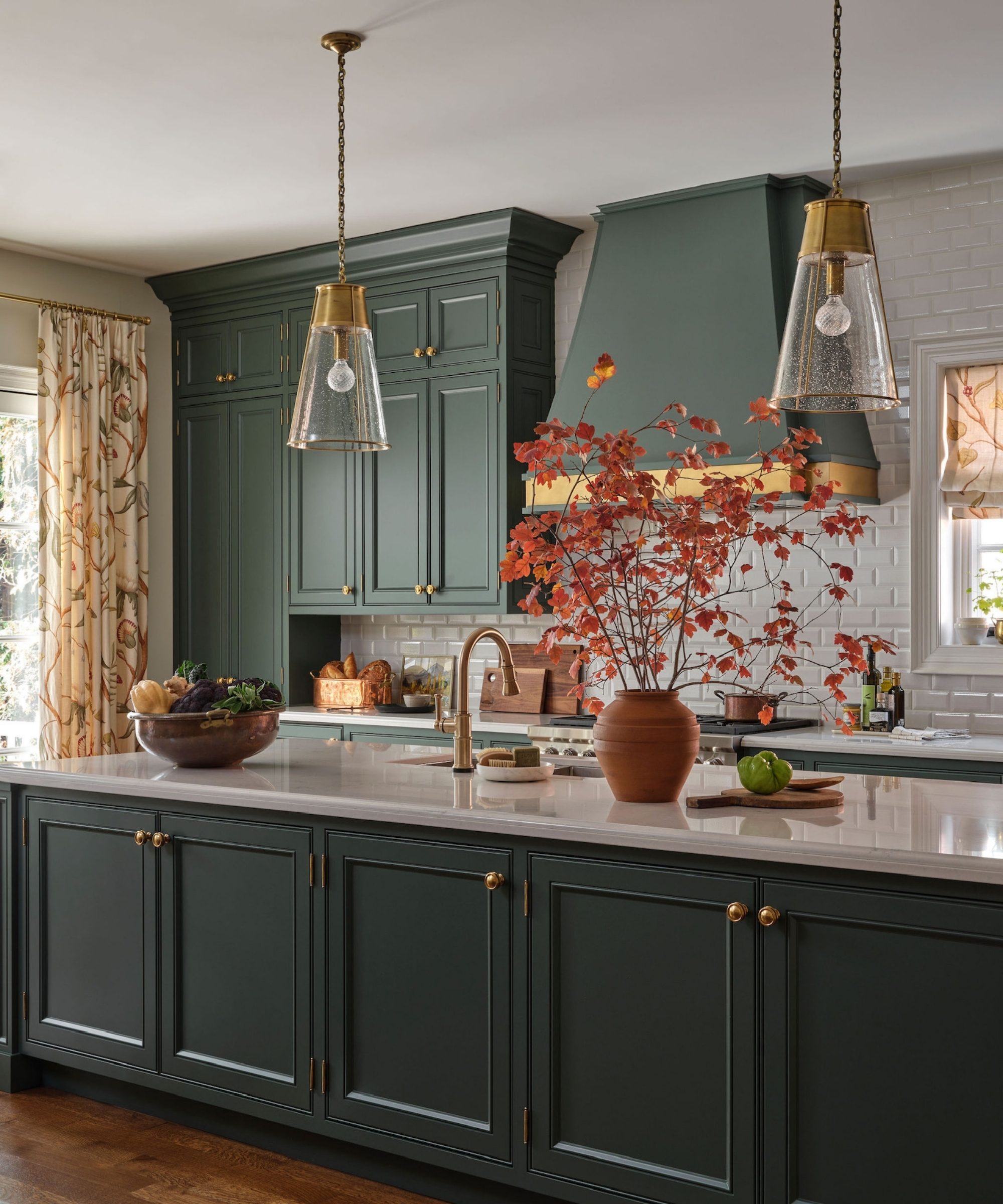 Dark green kitchen with brass lighting, terracotta vase filled with red fall branches and a bowl of seasonal produce.