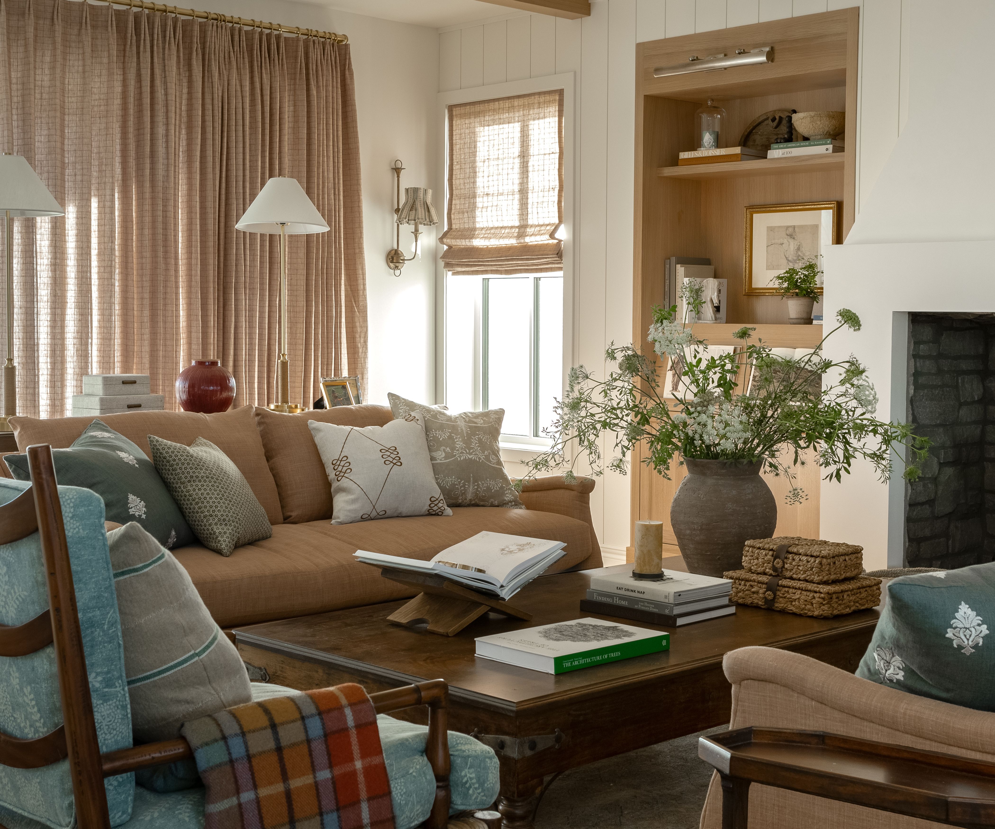 farmhouse meets grandma chic aesthetic white living room with tongue and groove walls, light oak built in shelving, fireplace, soft pink couch, skirted floral ottoman and two dark wood armchairs with blue cushions
