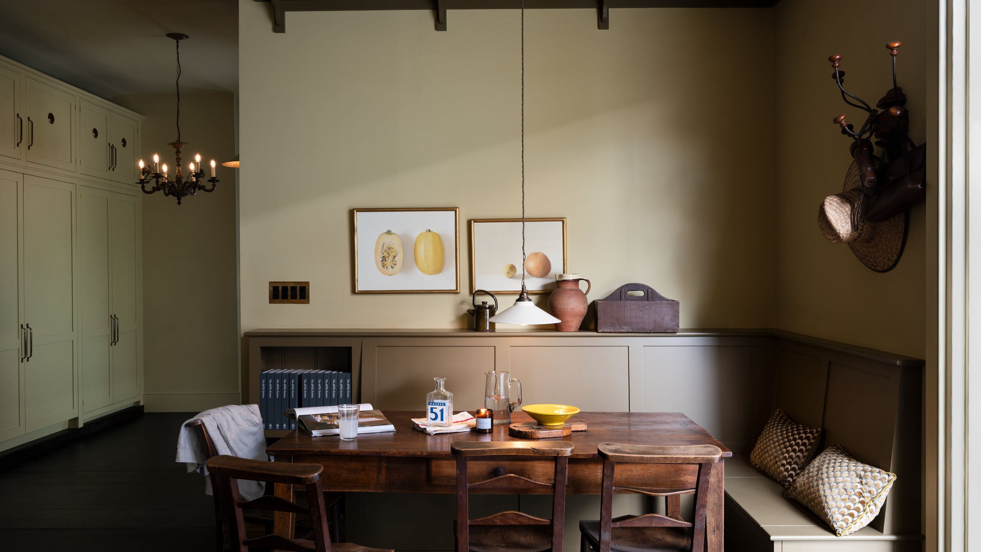 Shaker-style dining nook with sage green walls, dark wood dining table, built-in banquette seating, rustic ceramic pots, still life paintings of fruit on the walls, and wooden beams.