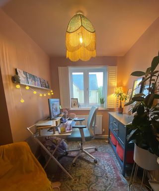 Side shot of small home office with terracotta colored walls and ceiling, green tasseled ceiling lampshade, white shutters, white desk, yellow chair and blue sideboard with plants and photos
