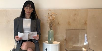 A young woman dressed in casual smart work attire sits on a transparent resin chair against a wood-paneled wall while holding a white pamphlet next to a cylindrical coffee table decorated with a water green glass vase.