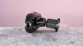 The Pitaka MagEZ Car Mount Pro 2 Qi2 sits on a stone-effect surface with a pink background. The view is of the base of the charging panel, revealing the USB-C port. The collar fasteners and the metal attachment hooks are also visible.