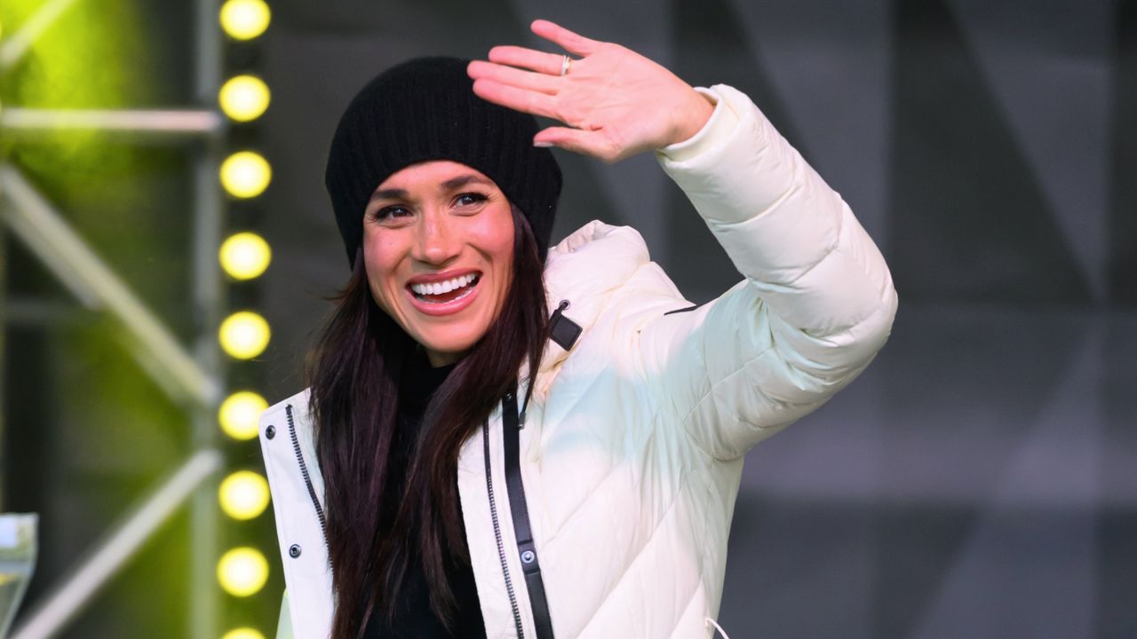 Meghan Markle waving in a white puffer jacket on stage with lights behind her 